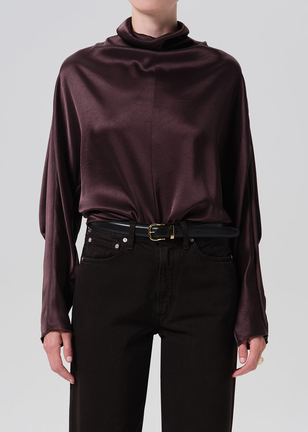 A model wearing a dark plum satin long-sleeve top with a cowl neck, tucked into black jeans with a black belt.