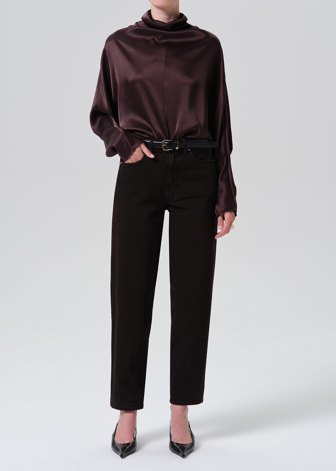 A model wearing a dark plum satin cowl-neck blouse tucked into black cropped trousers with a thin black belt, paired with black pointed-toe heels, against a grey background.