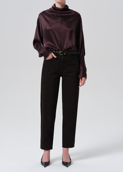 A model wearing a dark plum satin cowl-neck blouse tucked into black cropped trousers with a thin black belt, paired with black pointed-toe heels, against a grey background.