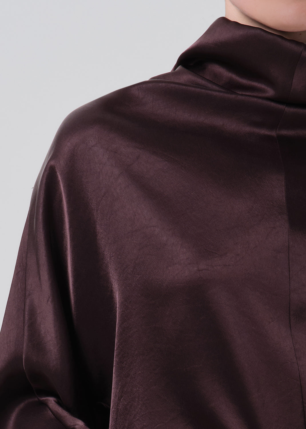 A close-up of a person wearing a dark plum satin-like top with a high mock neck, showing the fabric's soft sheen and texture against a grey background.