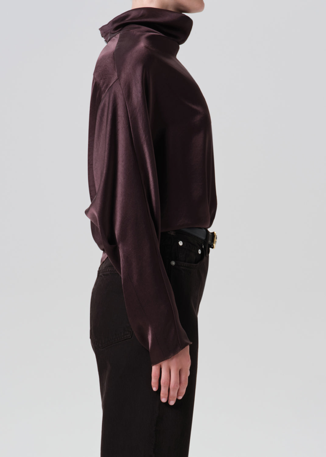 A side-profile view of a model wearing a dark plum satin turtleneck top and black pants against a grey background.