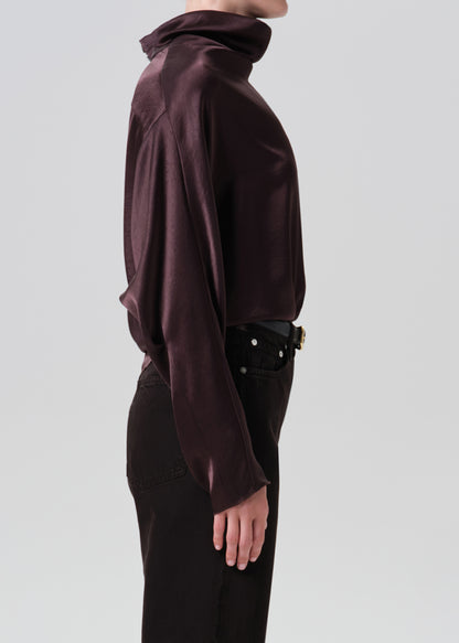 A side-profile view of a model wearing a dark plum satin turtleneck top and black pants against a grey background.