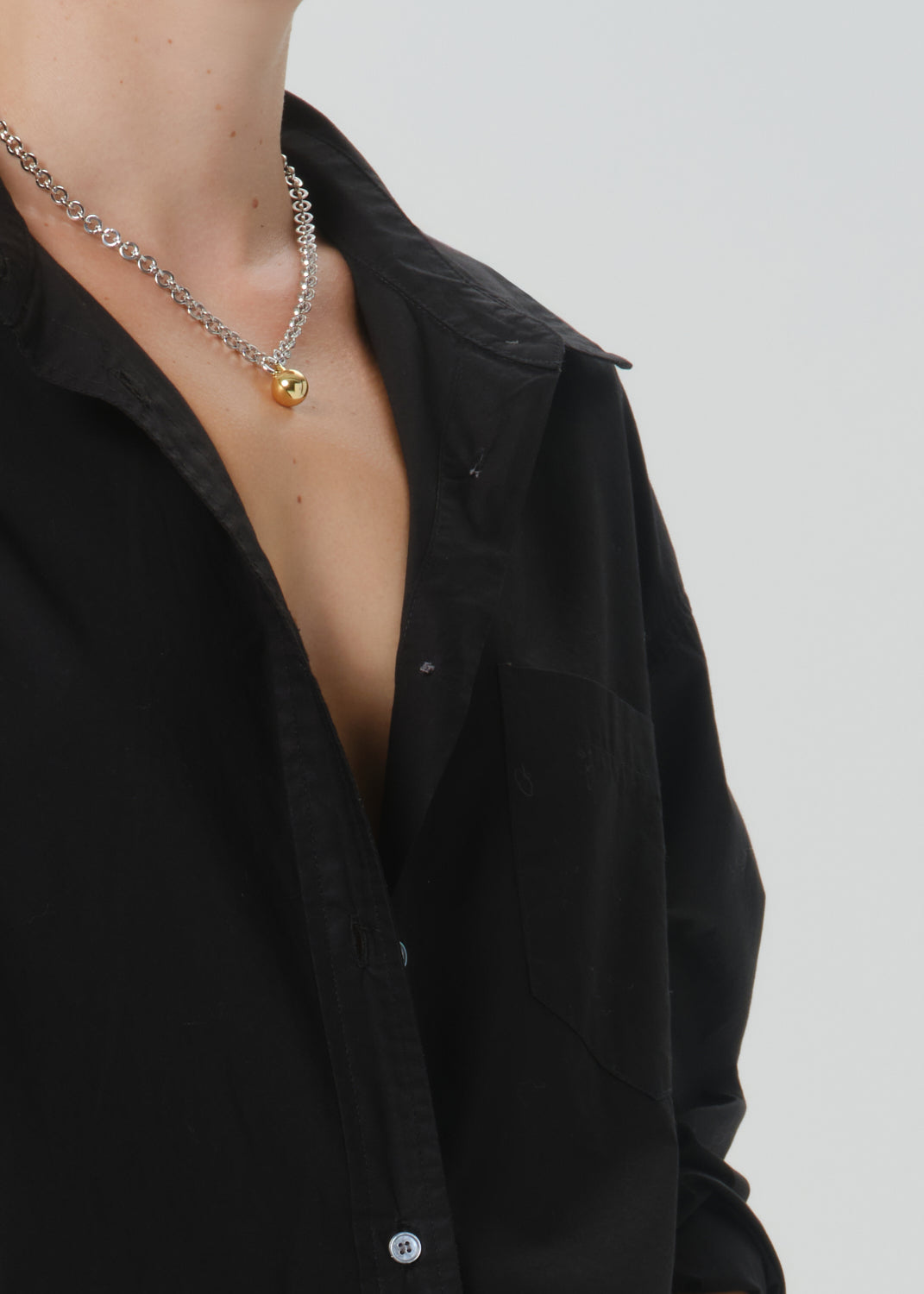 A close-up of a person wearing a silver chain necklace with a small gold pendant, paired with an open black button-up shirt grey background.