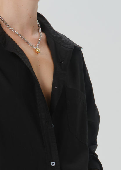 A close-up of a person wearing a silver chain necklace with a small gold pendant, paired with an open black button-up shirt grey background.