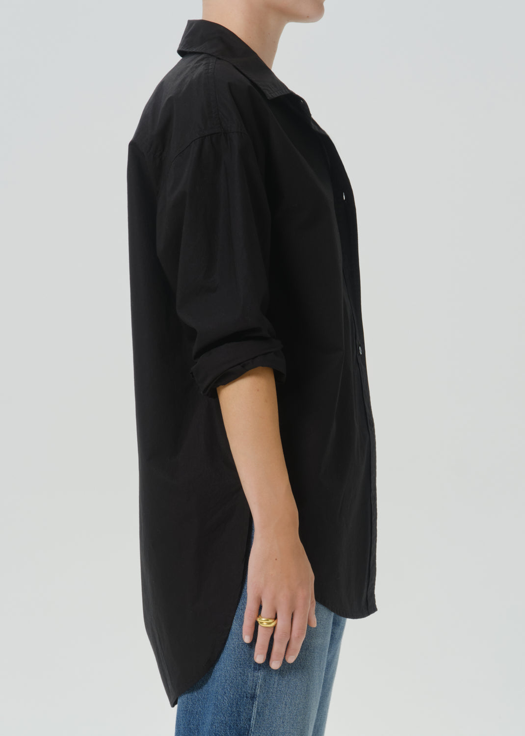 A side-profile view of a person wearing an oversized black button-up shirt with rolled sleeves and blue jeans against a grey background.