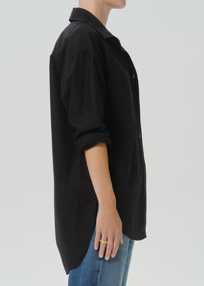 A side-profile view of a person wearing an oversized black button-up shirt with rolled sleeves and blue jeans against a grey background.