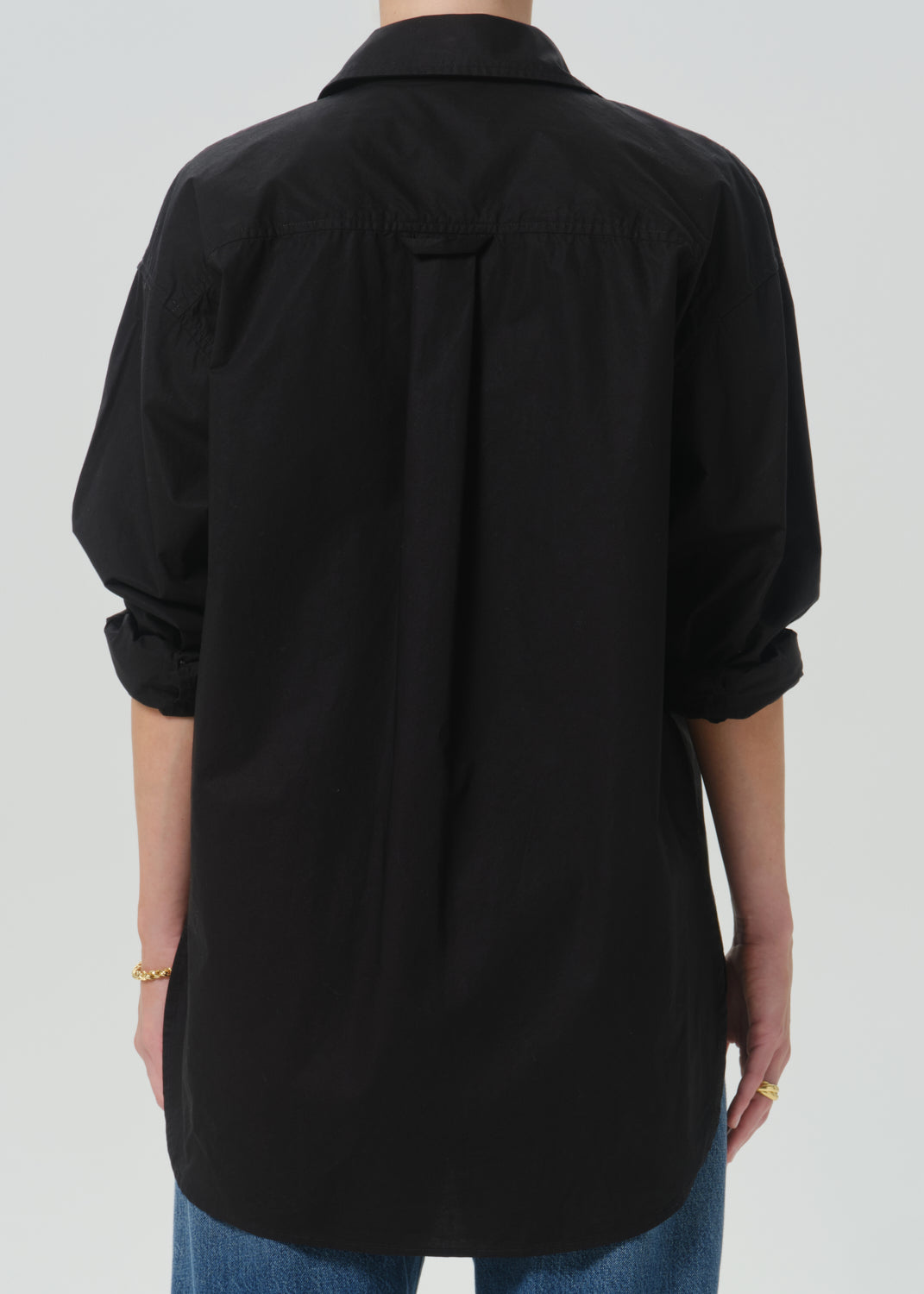 A person shown from the back wearing a black long-sleeve button-up shirt with rolled sleeves and blue jeans against a grey background.
