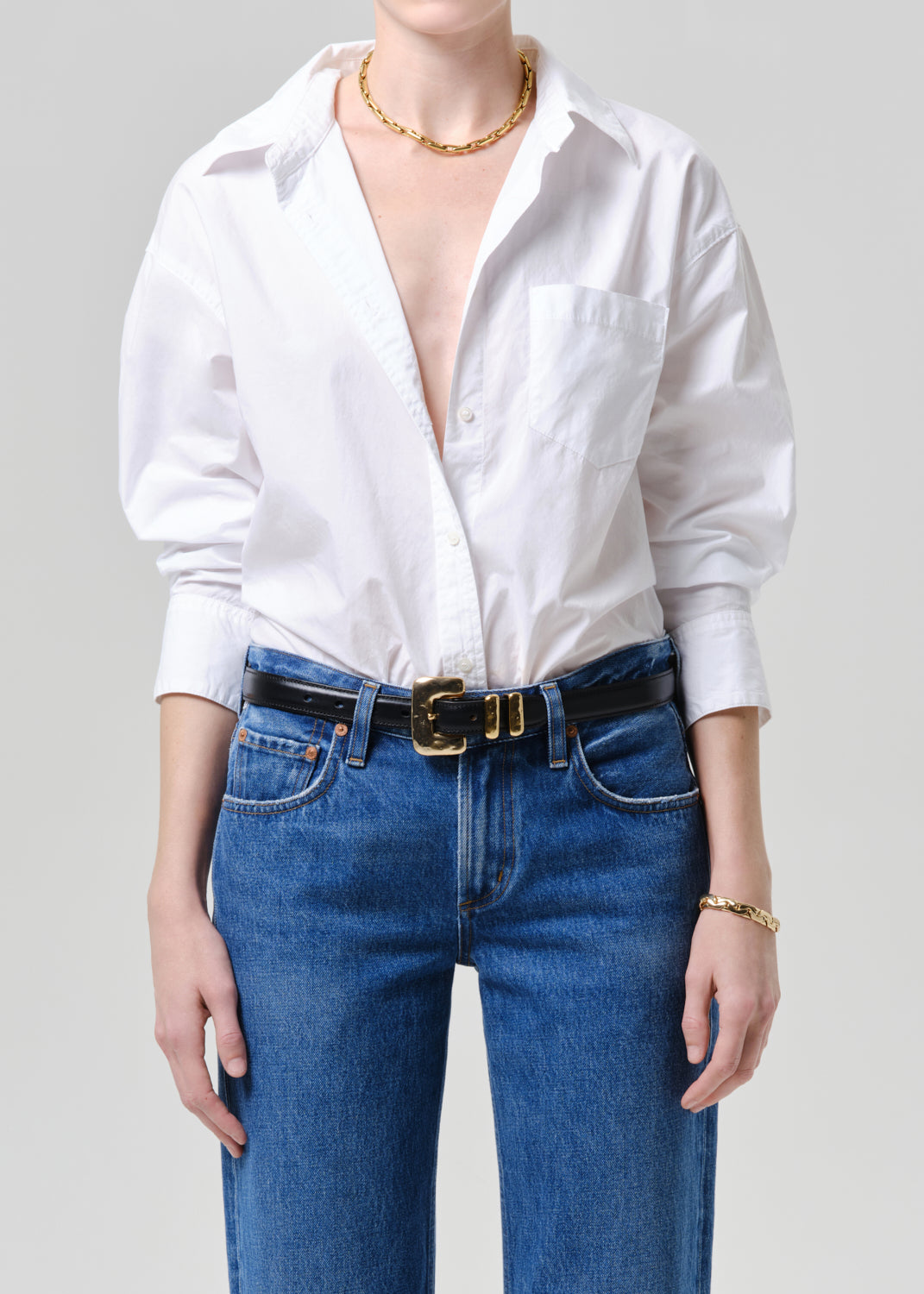 Person wearing a white blouse and blue jeans with a gold belt on a grey background