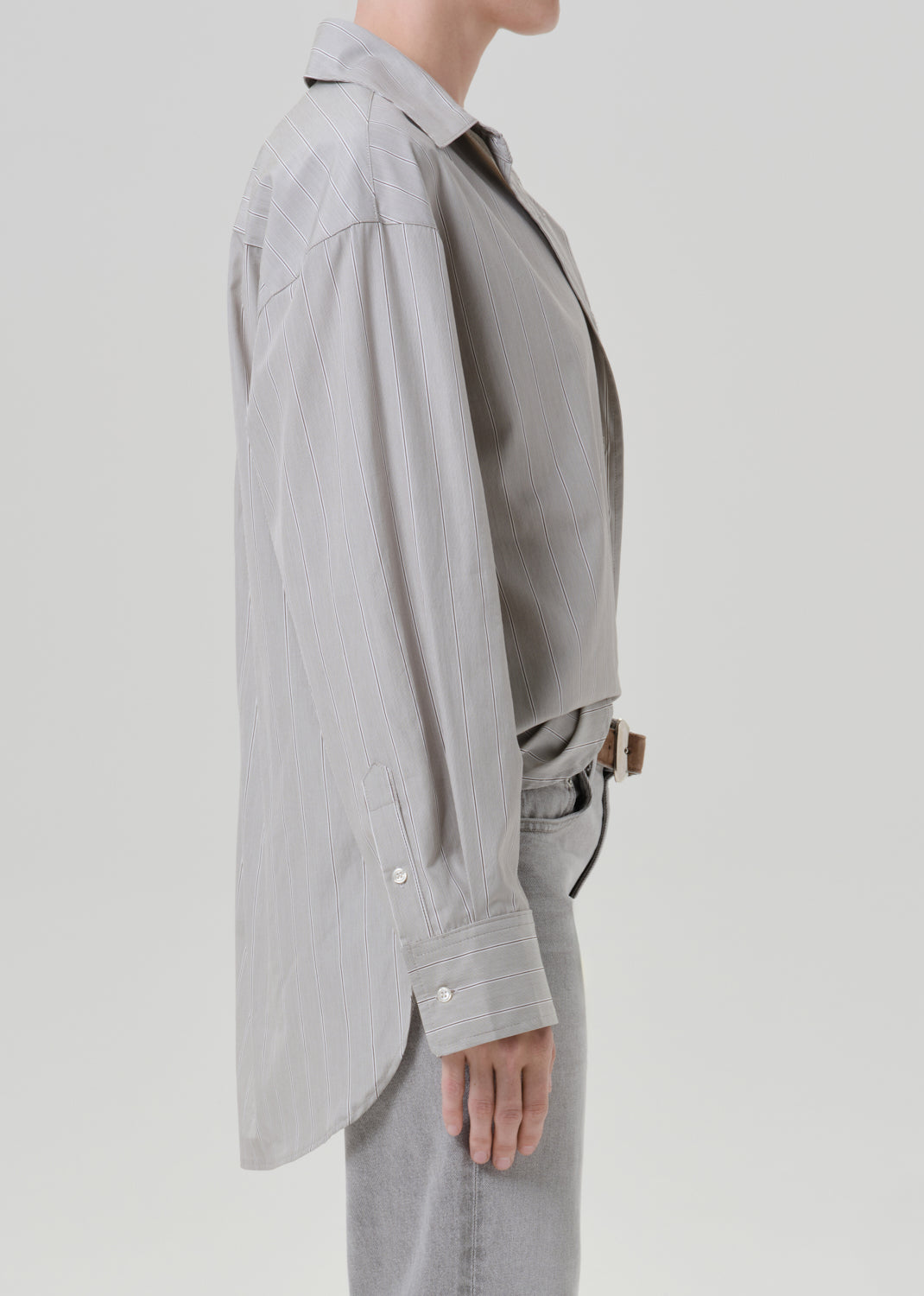 Person wearing a light grey striped button front blouse with light grey straight leg jeans viewed from the side on a grey background