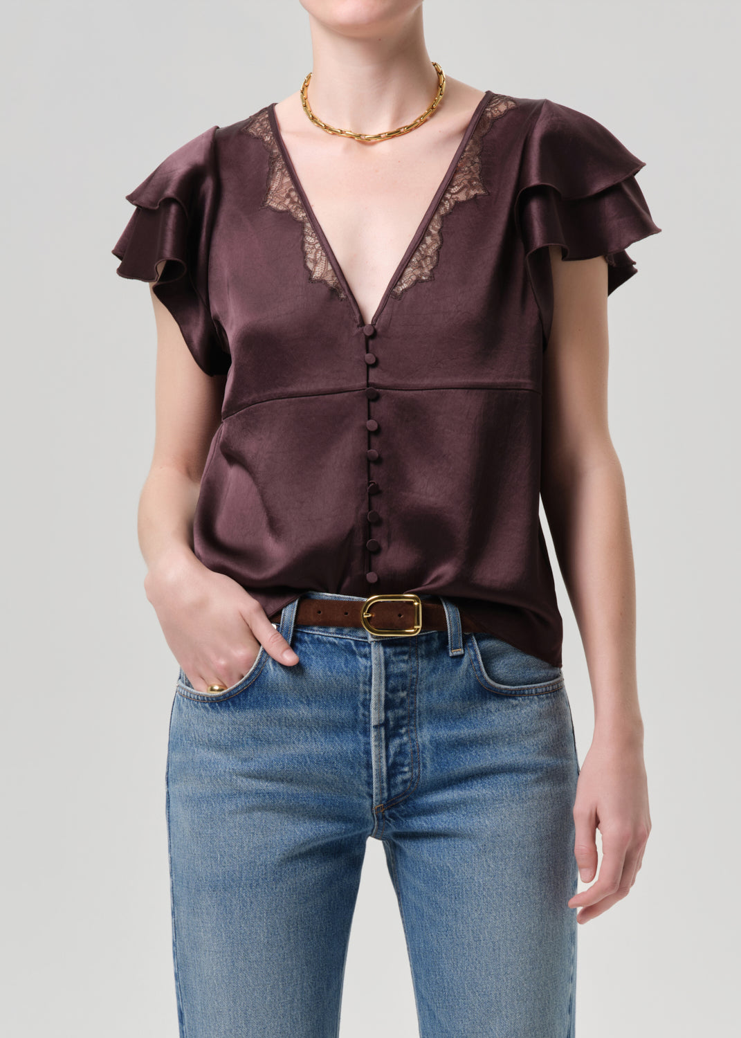 Woman wearing a brown satin top with ruffled sleeves and blue jeans on a grey background