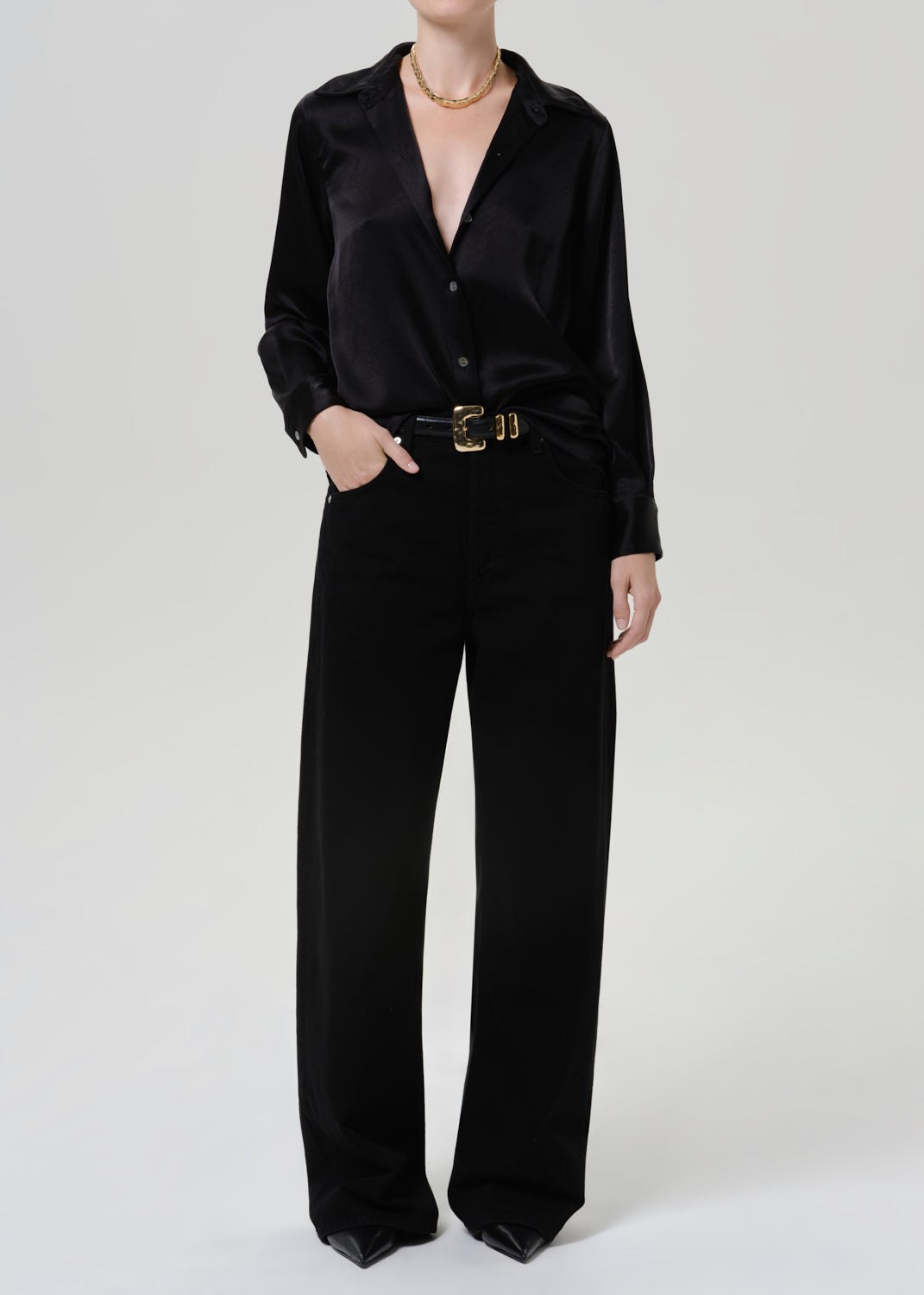 Woman wearing a black satin button-up shirt with black wide leg pants on a grey background.