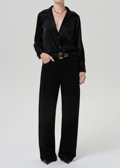 Woman wearing a black satin button-up shirt with black wide leg pants on a grey background.