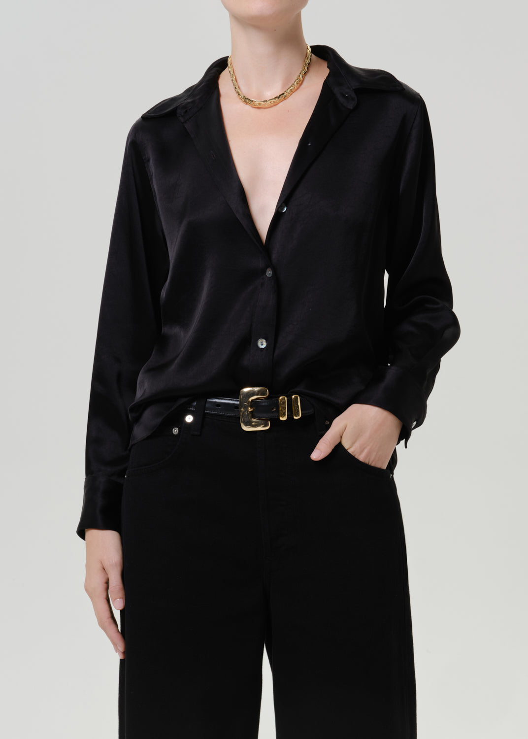 Woman wearing a black satin button-up shirt with black wide leg pants on a grey background.