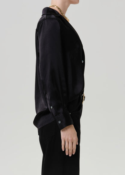Woman wearing a black satin button-up shirt with black wide leg pants viewed from the side on a grey background.
