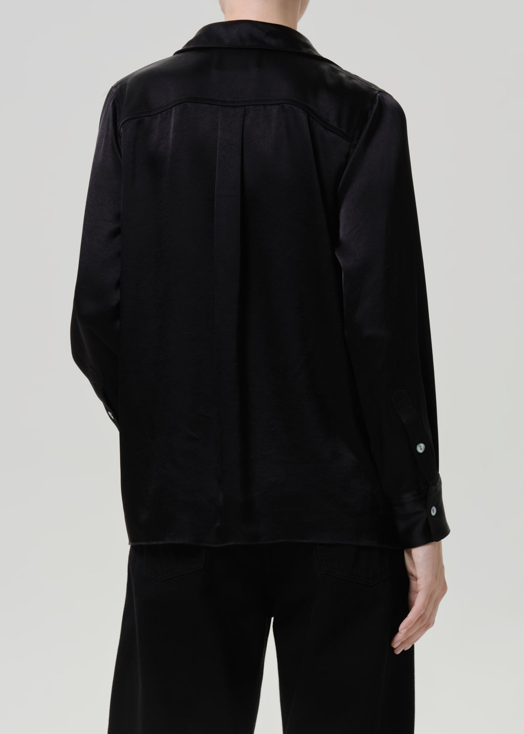 Woman wearing a black satin button-up shirt with black wide leg pants viewed from the back on a grey background.