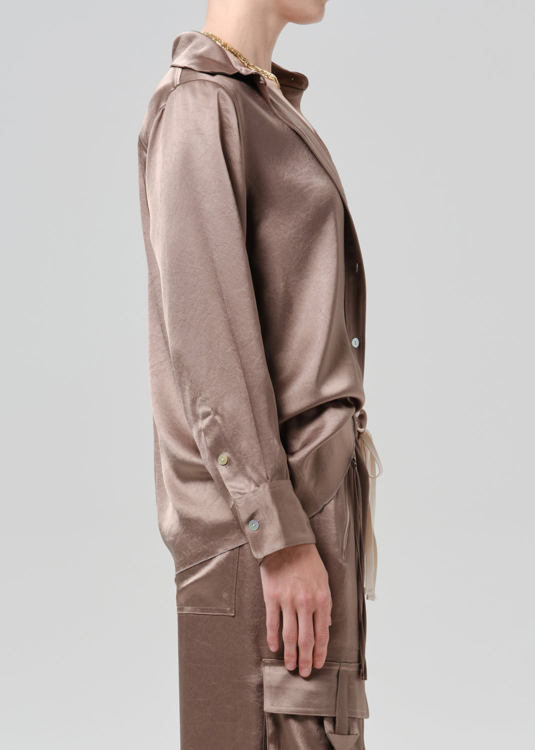 Person wearing a taupe satin button front blouse with taupe cargo pants viewed from the side on a grey background