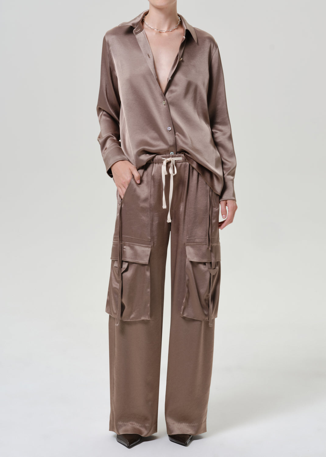 Woman wearing a taupe satin button-up shirt with matching taupe pants on a grey background.