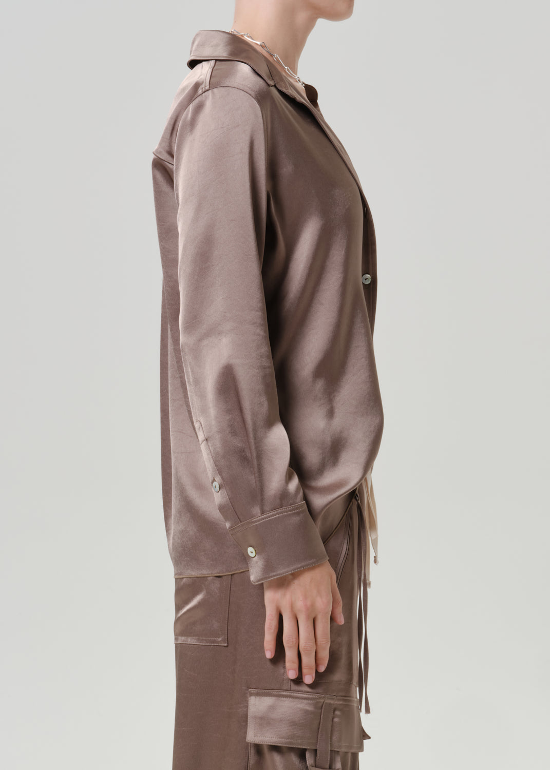 Woman wearing a taupe satin button-up shirt with matching taupe pants viewed from the side on a grey background.