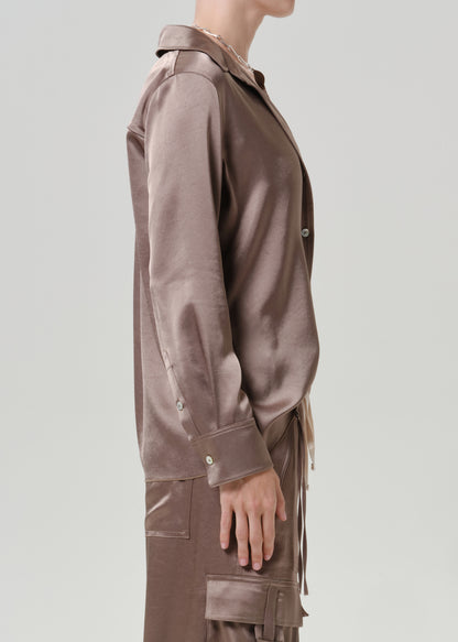 Woman wearing a taupe satin button-up shirt with matching taupe pants viewed from the side on a grey background.