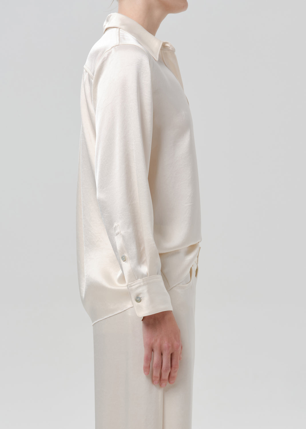 Person wearing an ivory satin button front blouse with ivory wide leg trousers viewed from the side on a grey background