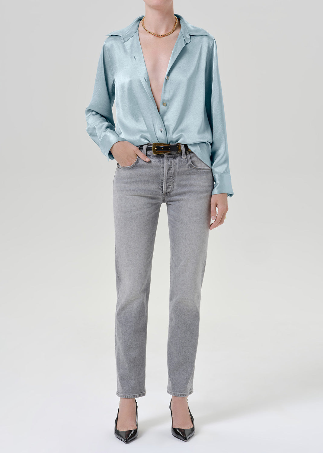 Woman wearing a light blue satin button-up shirt with grey straight leg jeans on a grey background.