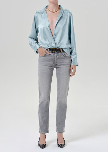 Woman wearing a light blue satin button-up shirt with grey straight leg jeans on a grey background.