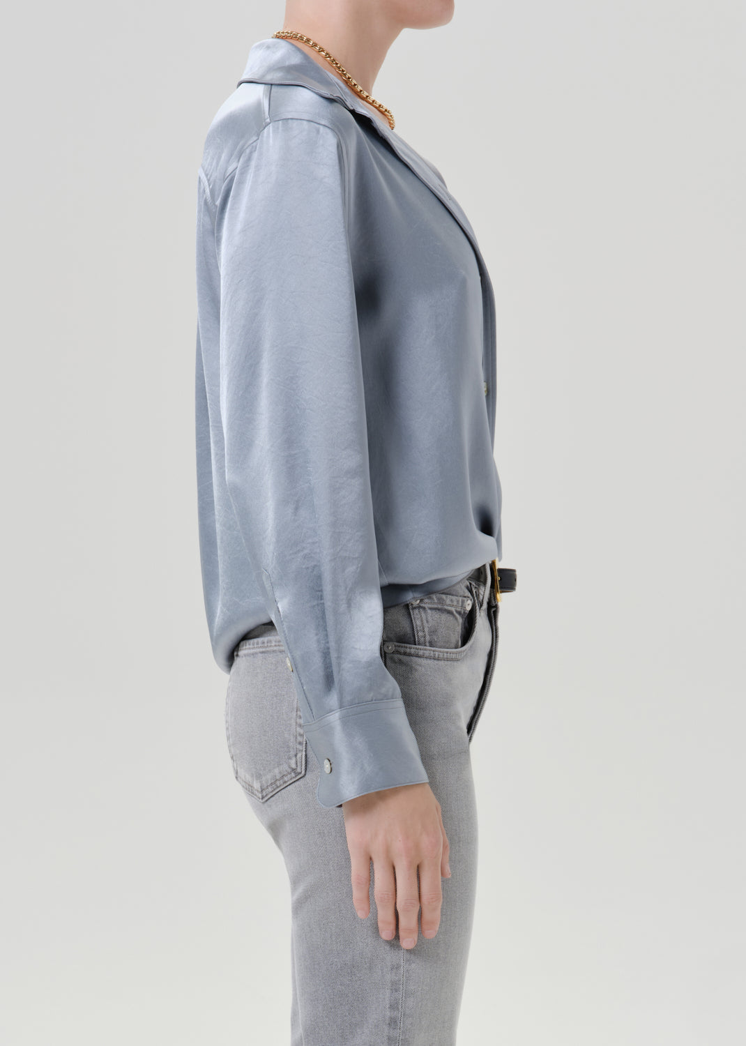 Person wearing a light blue satin button front blouse with light grey straight leg jeans viewed from the side on a grey background