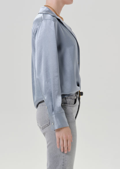 Person wearing a light blue satin button front blouse with light grey straight leg jeans viewed from the side on a grey background