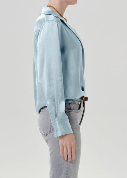 Woman wearing a light blue satin button-up shirt with grey straight leg jeans viewed from the side on a grey background.