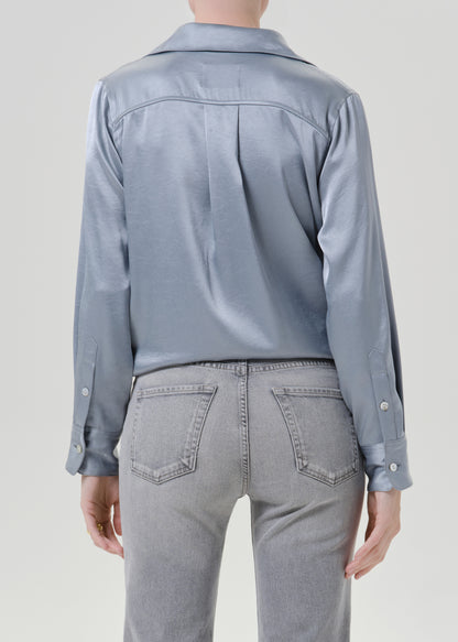 Person wearing a light blue satin button front blouse with light grey straight leg jeans viewed from the back on a grey background