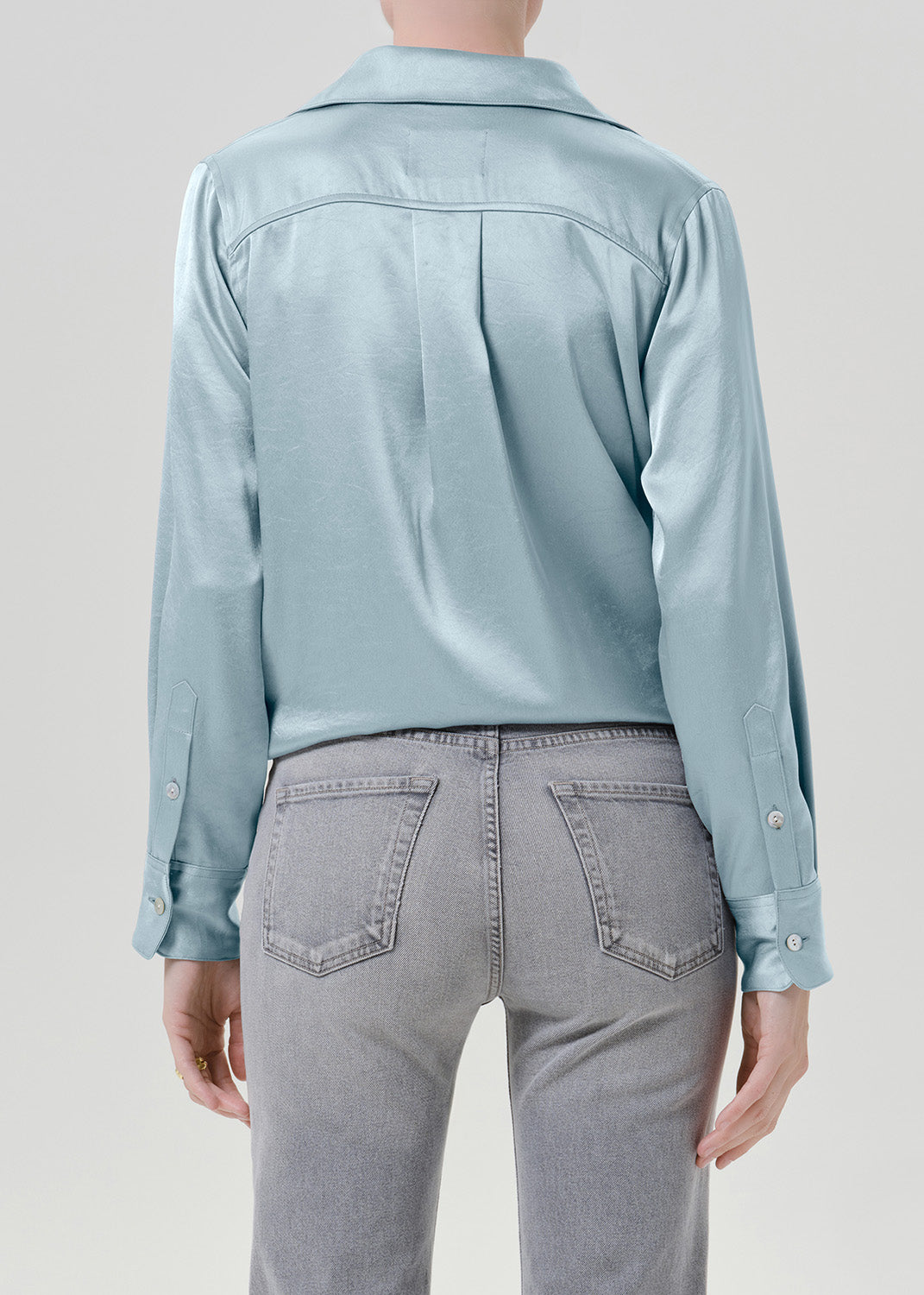 Woman wearing a light blue satin button-up shirt with grey straight leg jeans viewed from the back on a grey background.
