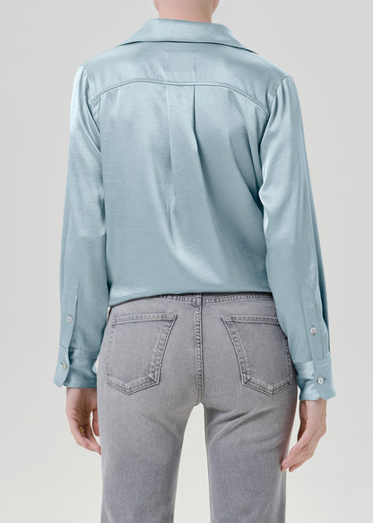 Woman wearing a light blue satin button-up shirt with grey straight leg jeans viewed from the back on a grey background.