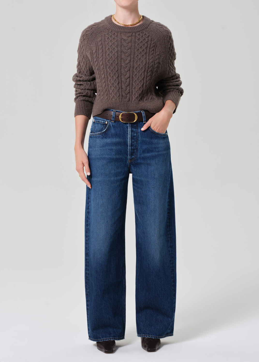 Person wearing a brown cable knit sweater and blue jeans on a white background