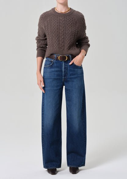 Person wearing a brown cable knit sweater and blue jeans on a white background