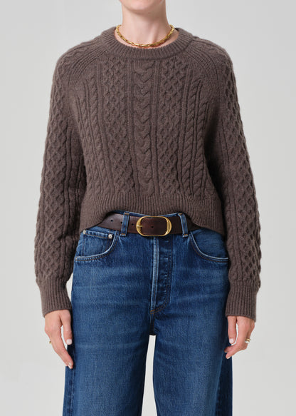 Person wearing a brown cable knit sweater and blue jeans on a plain background