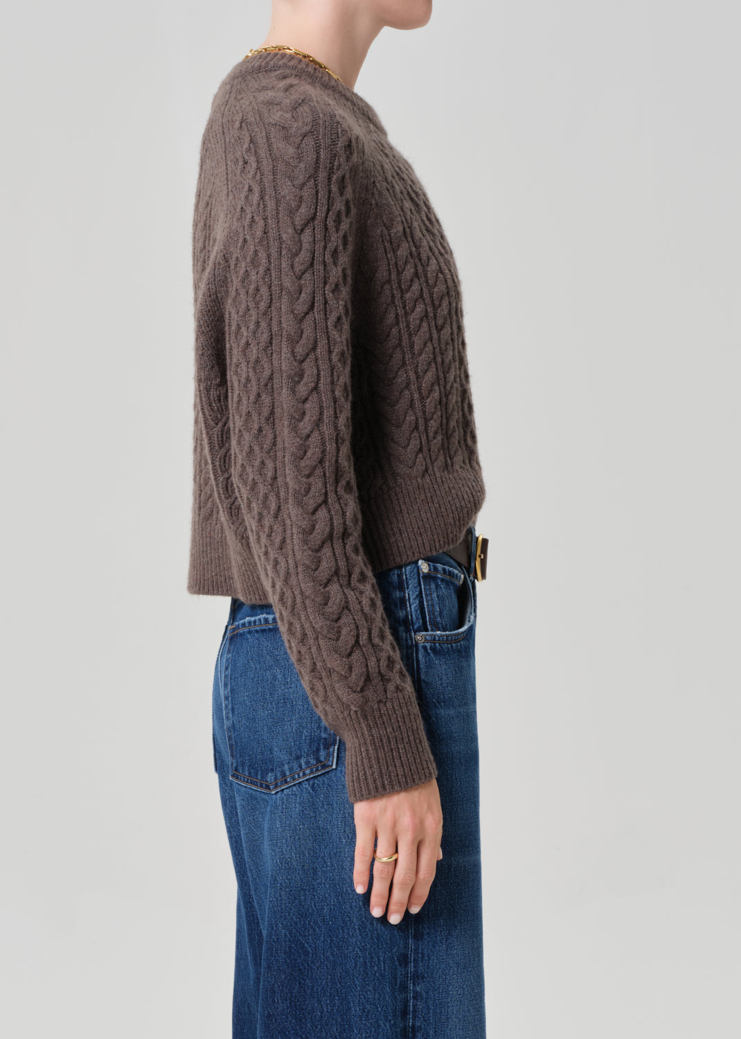 Person wearing a brown cable knit sweater and blue jeans on a light gray background