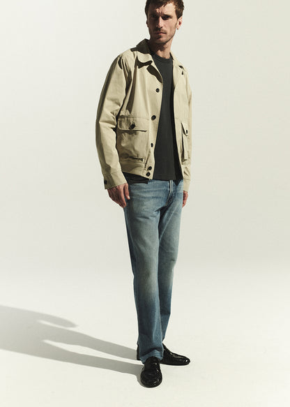 Man wearing a beige jacket, grey shirt, and blue jeans on a white background