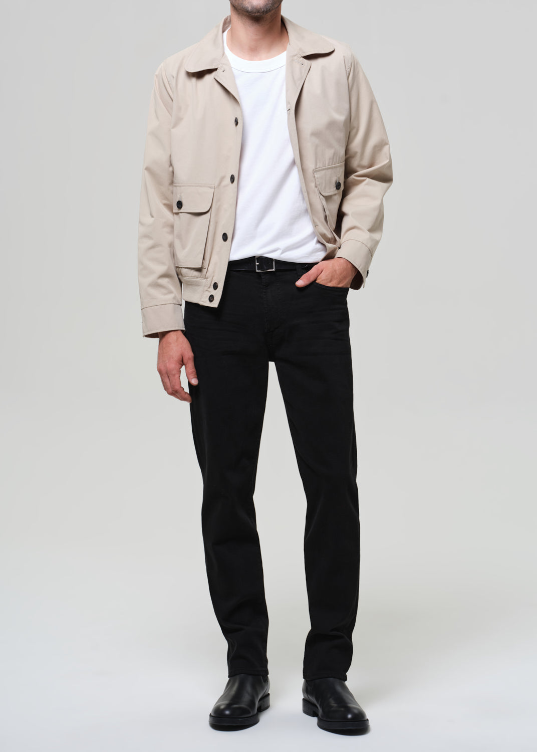 Man wearing a beige jacket, white shirt, black pants, and black shoes on a light gray background