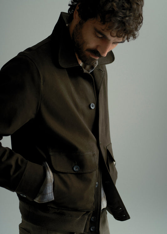 A man wearing a dark brown utility jacket over a light-colored shirt, looking down against a plain light gray background.