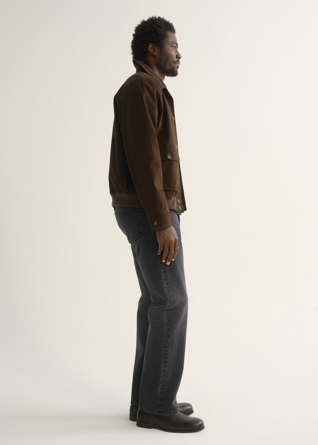 Person wearing a dark brown jacket with charcoal grey jeans and black shoes, side full body view on a light grey background.