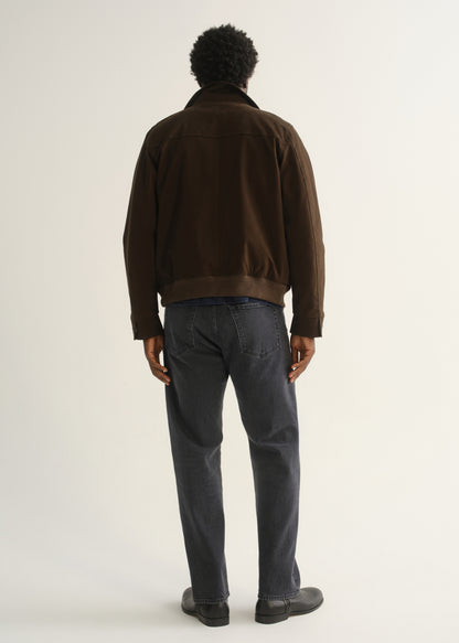 Person wearing a dark brown jacket with charcoal grey jeans and black shoes, back full body view on a light grey background.