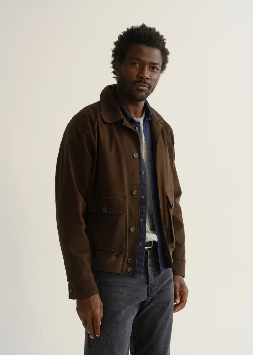 Person wearing a dark brown jacket over a blue plaid shirt with charcoal grey jeans, front cropped view on a light grey background.