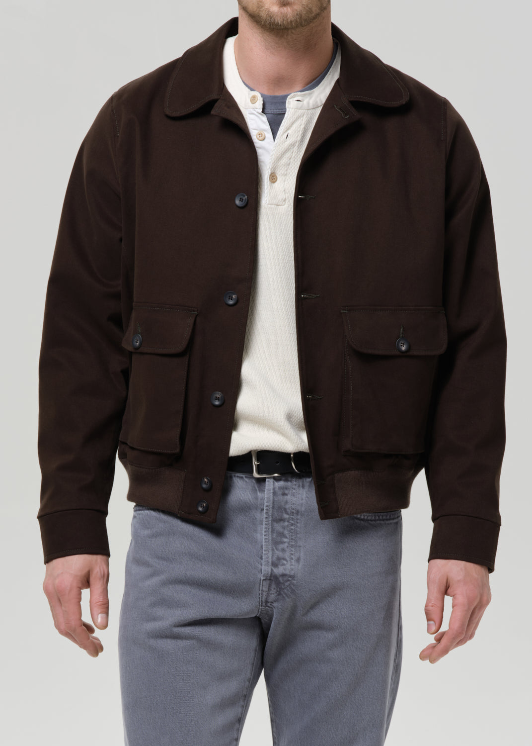 Man wearing a olive jacket over a white shirt with grey pants on a grey background