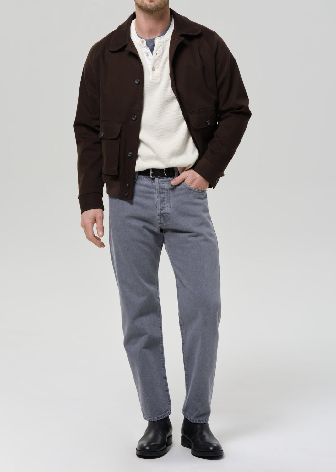 Man wearing a olive jacket, white shirt, grey pants, and black shoes on a grey background