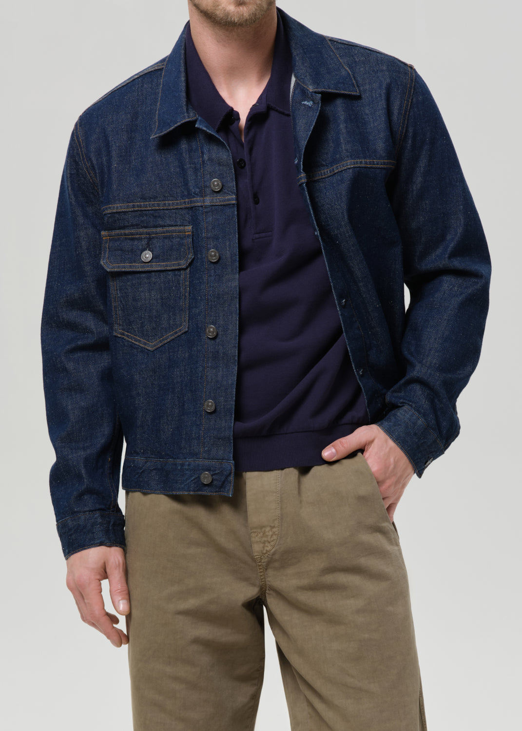 A man wearing a dark blue denim jacket over a navy polo shirt and khaki pants, shown from the neck down against a grey background.