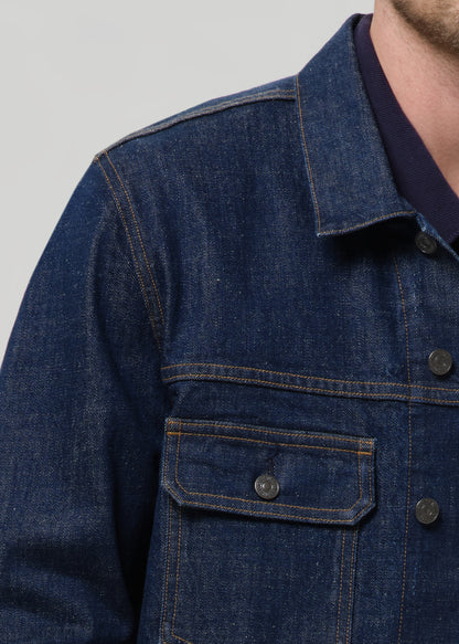 Close-up of a dark blue denim jacket with contrast stitching, a chest pocket, and a dark navy undershirt visible at the collar, on a grey background.