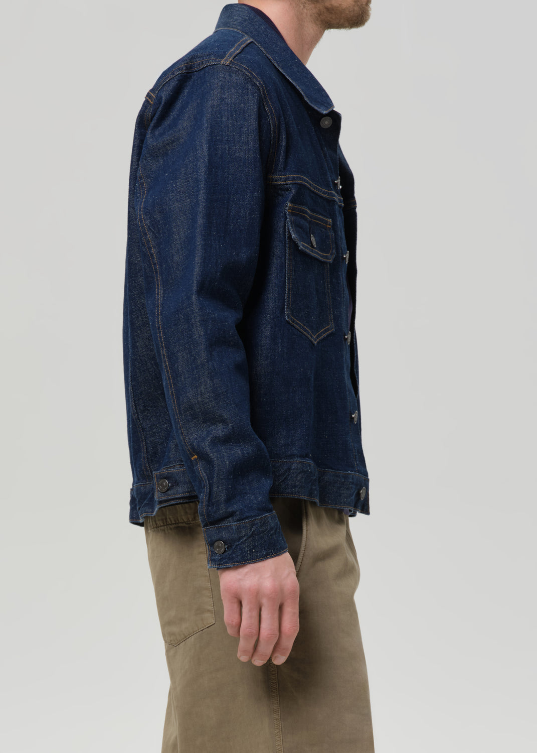 A man wearing a dark indigo blue denim jacket and khaki pants, shown from a side profile view against a grey background.