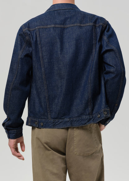 A person shown from the back wearing a dark indigo denim jacket and khaki pants against a grey background.