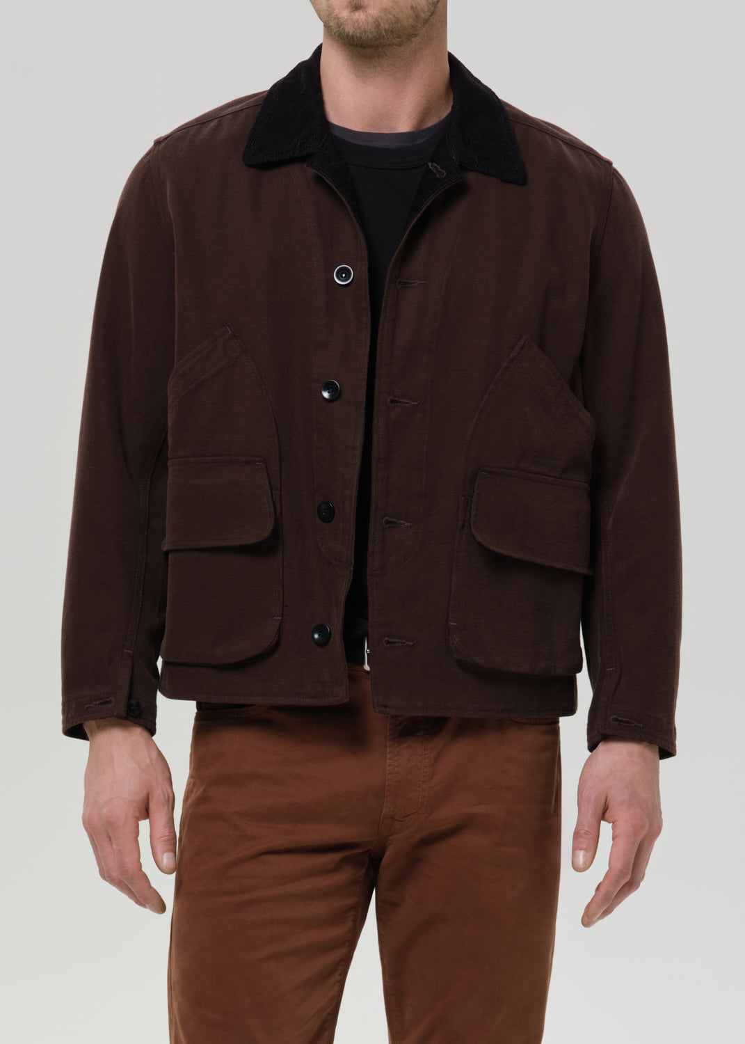 A man wearing a dark brown corduroy utility jacket with a black corduroy collar, layered over a black shirt and paired with brown pants, shot against a grey background.