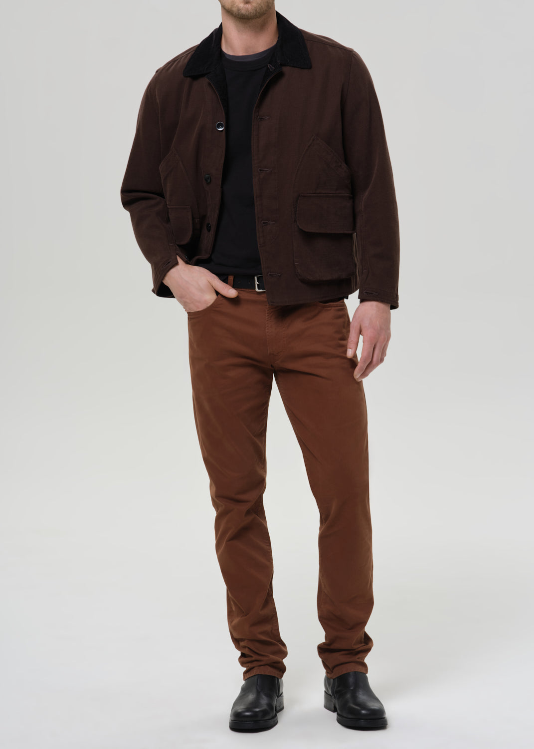 A man wearing a dark brown corduroy jacket over a black shirt and brown trousers, photographed against a grey background.
